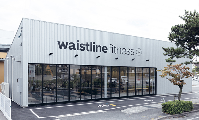 「waistline fitness」のロゴが映える、シルバーのガルバリウム外壁と大きなガラス窓が特徴的な店舗外観